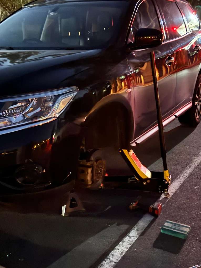 An SUV raised on a trolley jack and axle stand with its front wheel off, at night